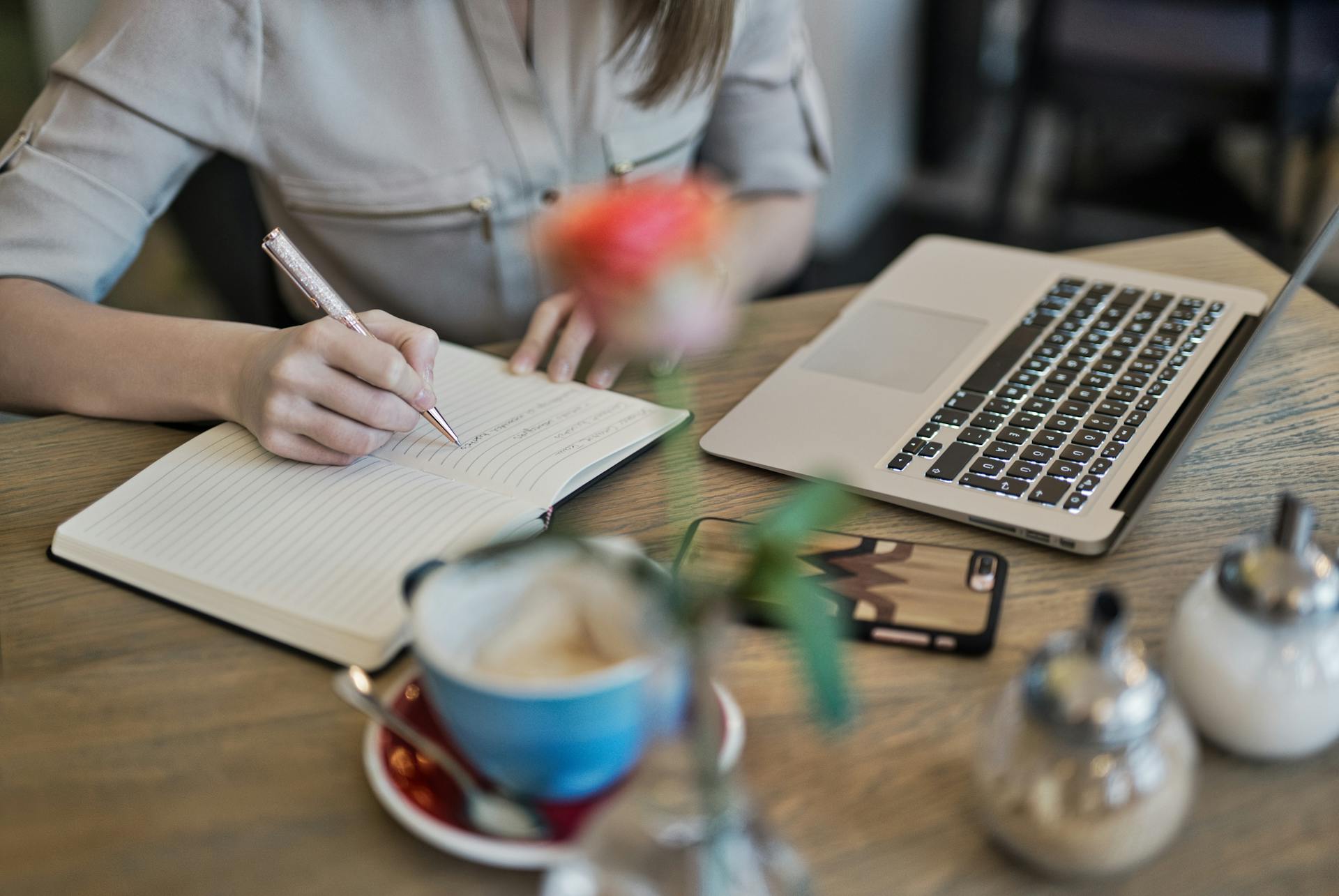 Foto von einem Schreibtisch an dem eine Person mit Notizblock, Laptop und Kaffee sitzt